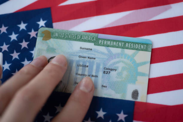 A hand holding a U.S. permanent resident card in front of a flag