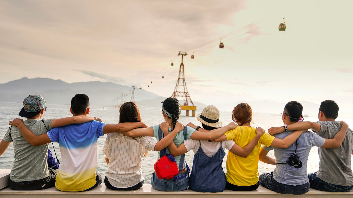 Group of young friends sitting on a wall with arms around shoulders, seen from behind
