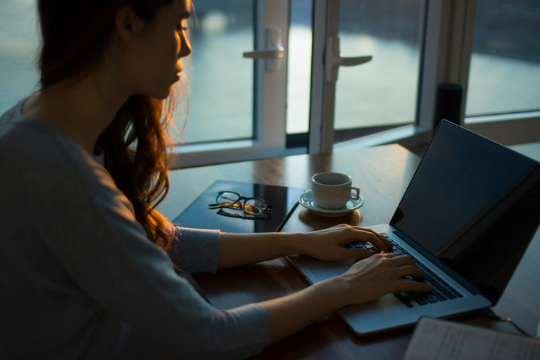 Woman typing on a laptop at a table by a window with a cup of coffee