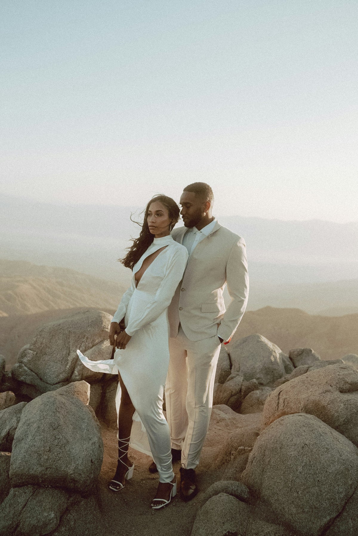 Couple in white wedding attire embracing on desert rocks at golden hour